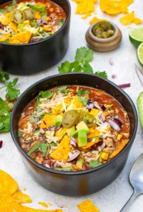 Two bowls of vegetarian taco soup topped with tortilla chips, avocado, red onion, grated cheese, coriander and jalapeños