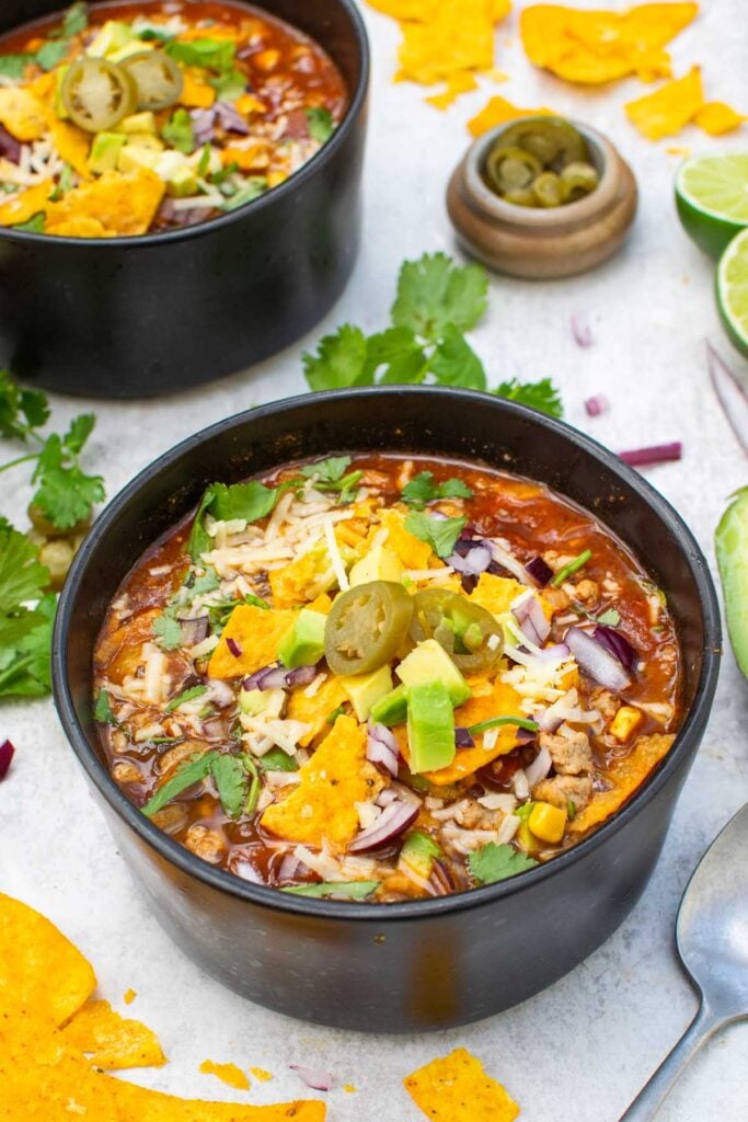 Two bowls of vegetarian taco soup topped with tortilla chips, avocado, red onion, grated cheese, coriander and jalapeños