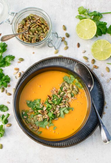 Thai pumpkin soup in a dark bowl, topped with toasted pumpkin and sunflower seeds and fresh coriander, next to a jar of pumpkin seeds and halved limes