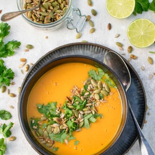 Thai pumpkin soup in a dark bowl, topped with toasted pumpkin and sunflower seeds and fresh coriander, next to a jar of pumpkin seeds and halved limes