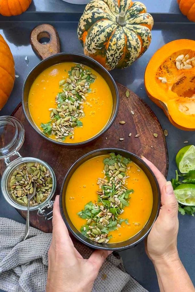 Hands holding a bowl of Thai pumpkin soup topped with toasted seeds and coriander, surrounded by whole and halved pumpkins, a jar of seeds and limes