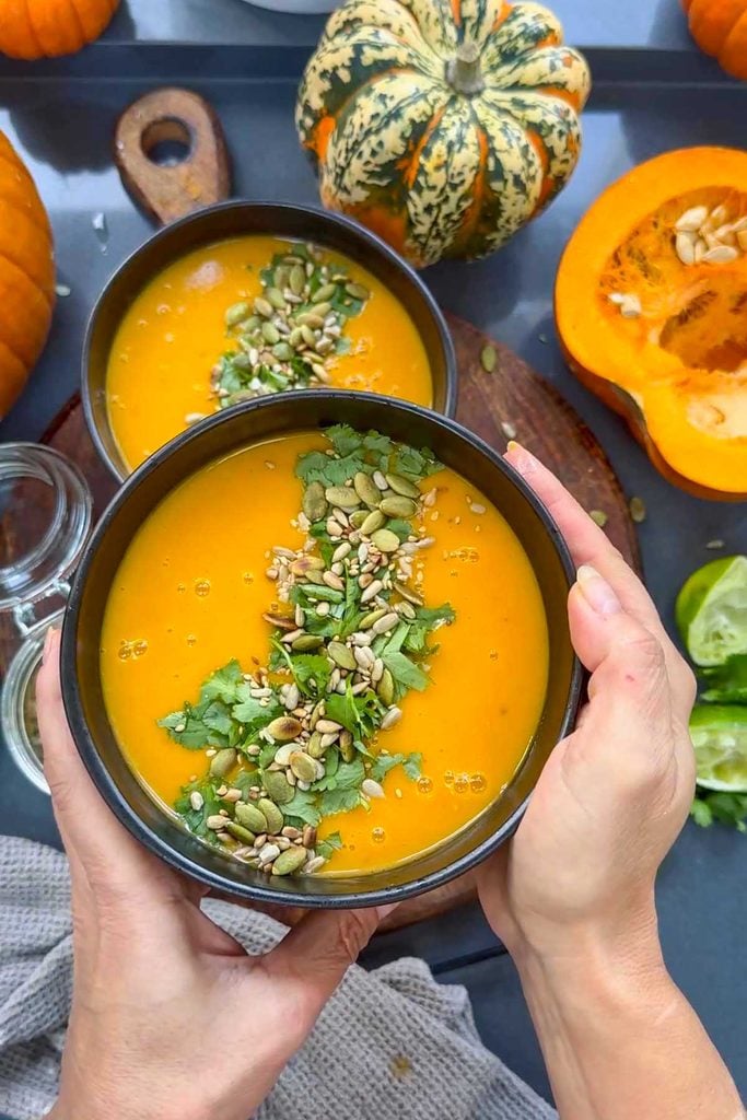Two bowls of vegan Thai pumpkin soup with seed and coriander topping, one held in both hands, on a wooden board with autumn squash