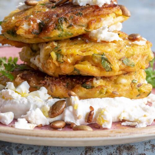 A stack of golden vegetable fritters on a bed of thick labneh, scattered with feta, pumpkin seeds and parsley