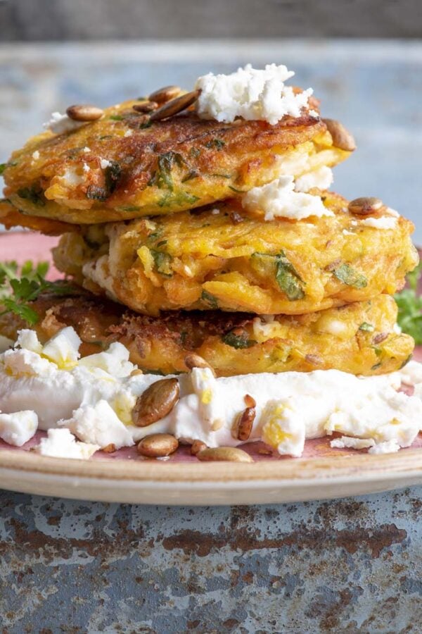 A stack of golden vegetable fritters on a bed of thick labneh, scattered with feta, pumpkin seeds and parsley